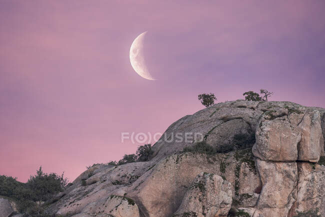 Bajo ángulo de espectacular paisaje de áspera montaña rocosa bajo el cielo rosado del atardecer con luna menguante en el Parque Nacional Sierra de Guadarrama - foto de stock