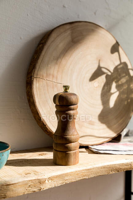 Molino de pimienta de madera y tablero redondo colocado en estante de madera rústica cerca de la pared en la cocina casera de estilo ecológico - foto de stock