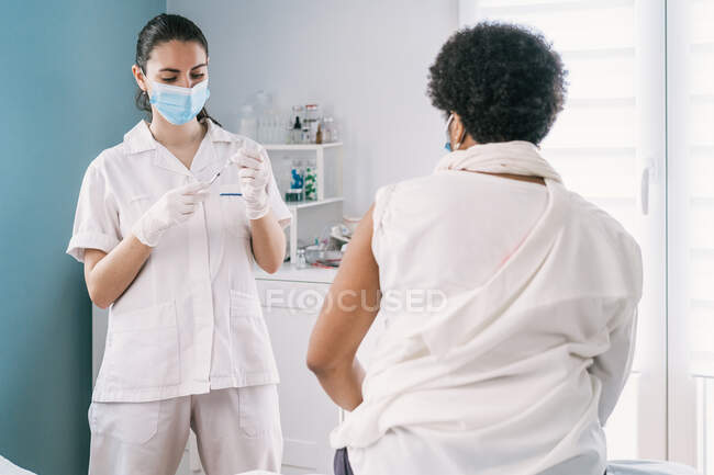 Medico donna in guanti di lattice e scudo facciale che riempie la siringa dal flacone con il vaccino che si prepara a vaccinare la paziente afroamericana matura non riconoscibile in clinica durante l'epidemia di coronavirus — Foto stock