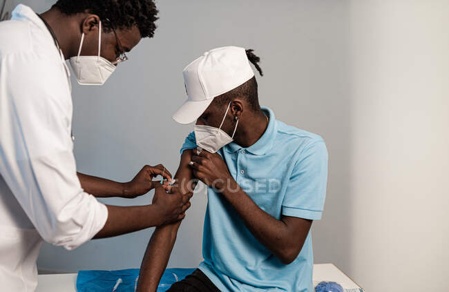 Чорний лікар в захисній формі і латексні рукавички вакцинують чоловічої афроамериканської пацієнтки в клініці під час спалаху коронавірусу. — стокове фото