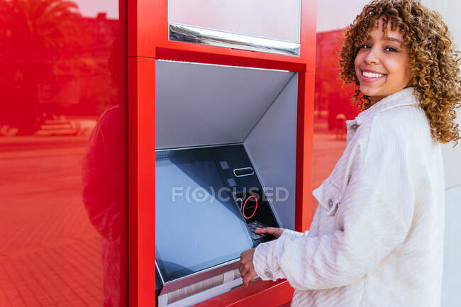 Side view of smiling African American female using ATM terminal and withdrawing cash while standing on city street looking at camera — Stock Photo
