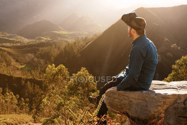 Seitenansicht eines männlichen Reisenden in VR-Brille, der mit virtueller Realität interagiert, während er bei Sonnenuntergang auf einem Hügel im bergigen Gelände sitzt — Stockfoto