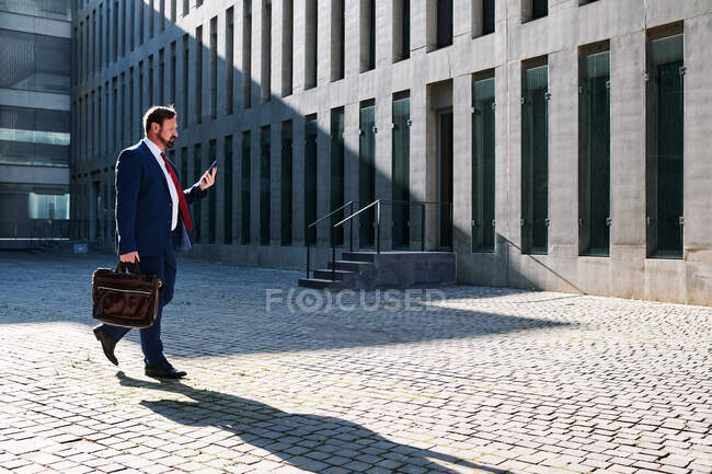 Впевнений чоловік-підприємець у елегантному костюмі, який ходить по вулиці в діловому районі і дивиться на мобільний телефон. — Stock Photo