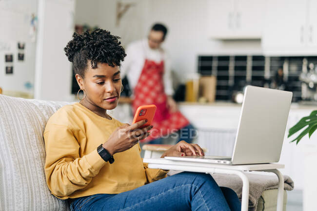 Hombre en delantal de navegación teléfono inteligente en la cocina y la mujer negra portátil de navegación y teléfono inteligente en casa - foto de stock