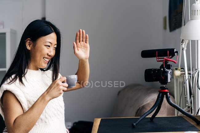 Sonriente blogger étnica con taza de bebida caliente mostrando un gesto de saludo mientras graba video en la cámara fotográfica en casa - foto de stock