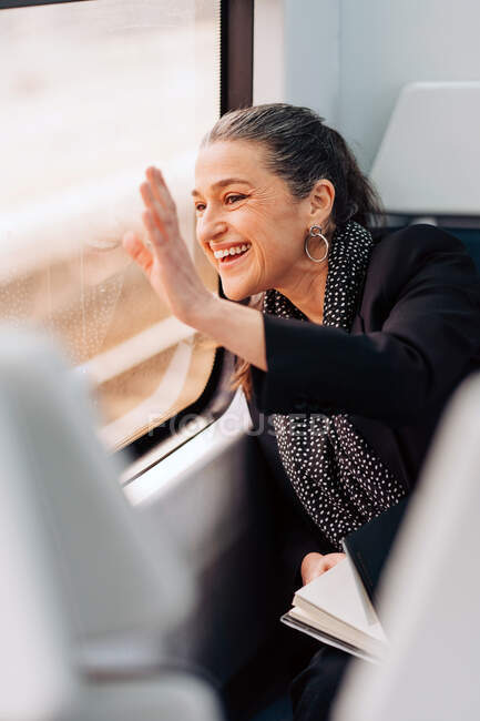 Side view of positive female waving hand and looking out window while sitting on passenger seat in wagon during ride — Stock Photo