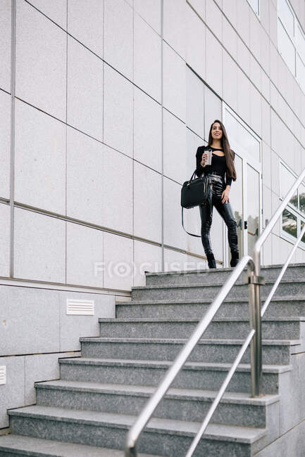 Low angle of trendy female entrepreneur with takeaway drink in cup standing near modern building on city and looking away — Stock Photo