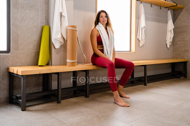 Tired glad sportswoman in sportswear sitting with towel on shoulders on wooden bench in gym locker room after training — Stock Photo