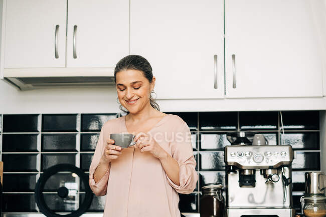 Весела жінка середнього віку з чашкою гарячого напою, що стоїть на кухонній стійці з білими шафами та сучасною кавовою машиною вдома — стокове фото