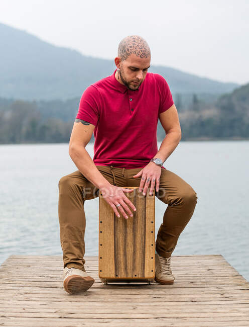 Homem percussionista concentrado sentado e jogando cajon no cais de madeira contra o rio calmo e montanhas em dia nublado — Fotografia de Stock