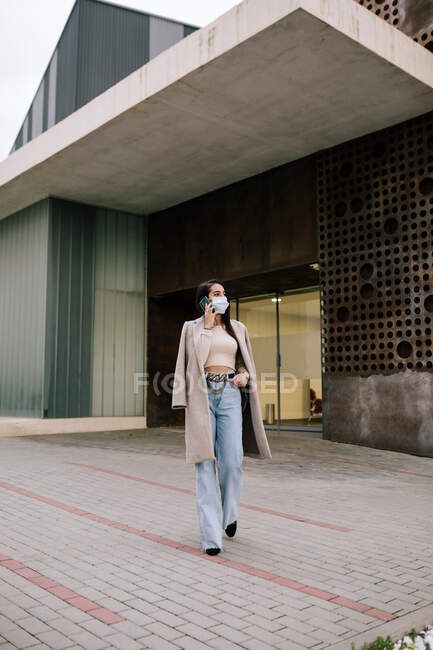 Mujer emprendedora de moda en ropa elegante caminando por la ciudad y hablando por teléfono móvil mientras mira hacia otro lado - foto de stock