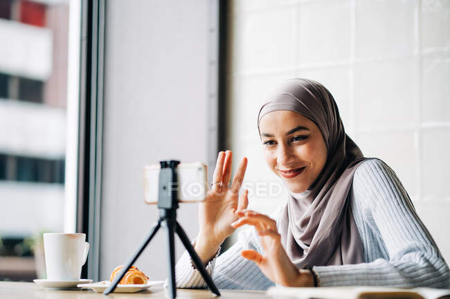 Low angle of cheerful Muslim female in headscarf having video call on smartphone on tripod for blog while sitting at table in cafe — Stock Photo