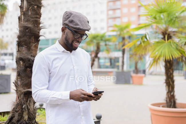 Hombre afroamericano de moda parado en la calle con palmeras y mensajería en las redes sociales a través del teléfono móvil - foto de stock
