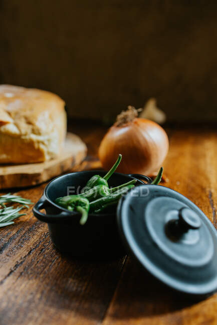 Pimenta fresca inteira em panela pequena perto de cebola crua e pão rústico na mesa de madeira — Fotografia de Stock