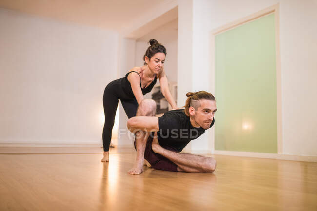 Barefoot female leaning forward while helping male friend stretching leg on shiny parquet in building — Stock Photo