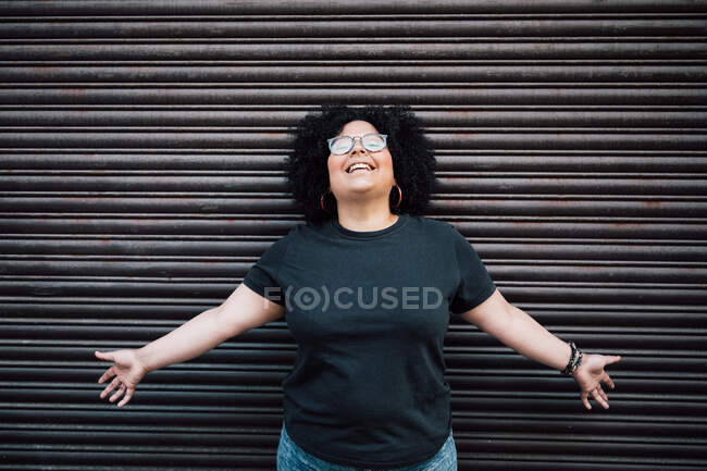 Conteúdo adulto com excesso de peso feminino em óculos e braços abertos cabelos encaracolados contra a parede com nervuras durante o dia — Fotografia de Stock