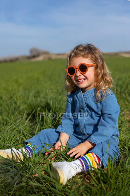 Cute happy little girl in trendy clothes and sunglasses sitting and relaxing on grassy lawn — Stock Photo