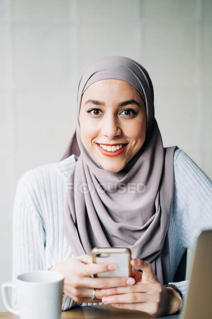 Ritratto di donna freelance etnica in hijab con smartphone seduta a tavola nel bar e che lavora a distanza — Foto stock