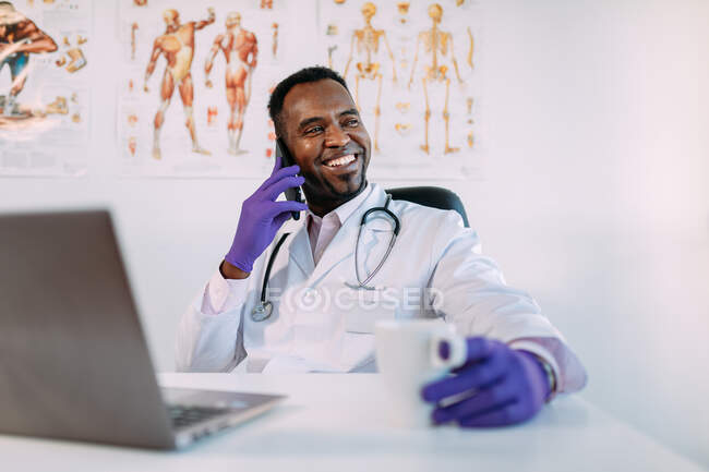 Joyeux jeune médecin afro-américain en vêtements médicaux et gants ayant une conversation téléphonique et souriant tout en étant assis à table avec ordinateur portable et tasse de café pendant la pause à la clinique — Photo de stock