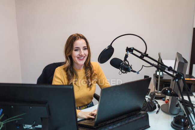 Позитивная женщина просматривает современный нетбук, сидя за столом с мониторами и микрофоном во время работы в студии вещания со специальным оборудованием — стоковое фото