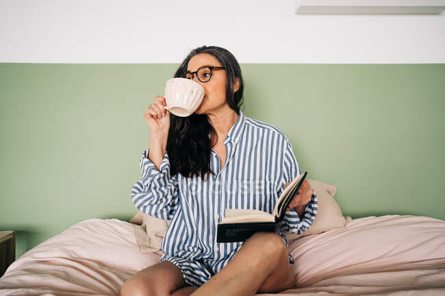 Femme intelligente dans des lunettes avec des cheveux longs et un cahier regardant loin et reposant sur un lit doux dans la chambre — Photo de stock