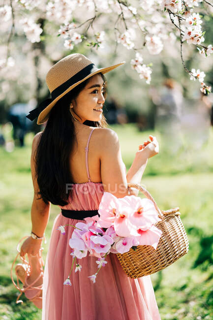 Назад вид босоніж жінка в сукні і солом'яному капелюсі стоїть з кошиком і взуттям у квітучому саду, дивлячись через плече — стокове фото