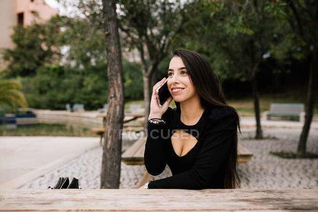 Усміхнена жінка-підприємець сидить за столом, розмовляючи на смартфоні, дивлячись далеко — стокове фото