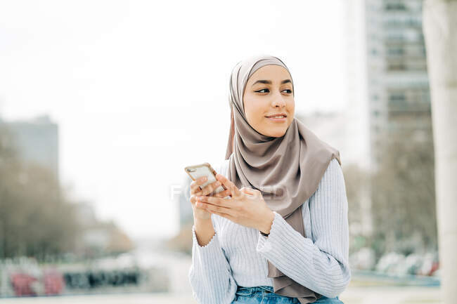 Young ethnic female in hijab standing in city and messaging on mobile phone — Stock Photo