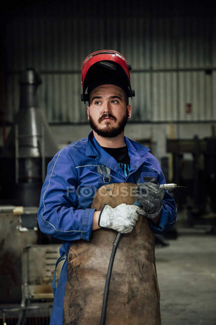 Mecânico em capacete de proteção e avental em azul de pé geral com mangueira de soldagem em oficina de luz perto de construções metálicas — Fotografia de Stock