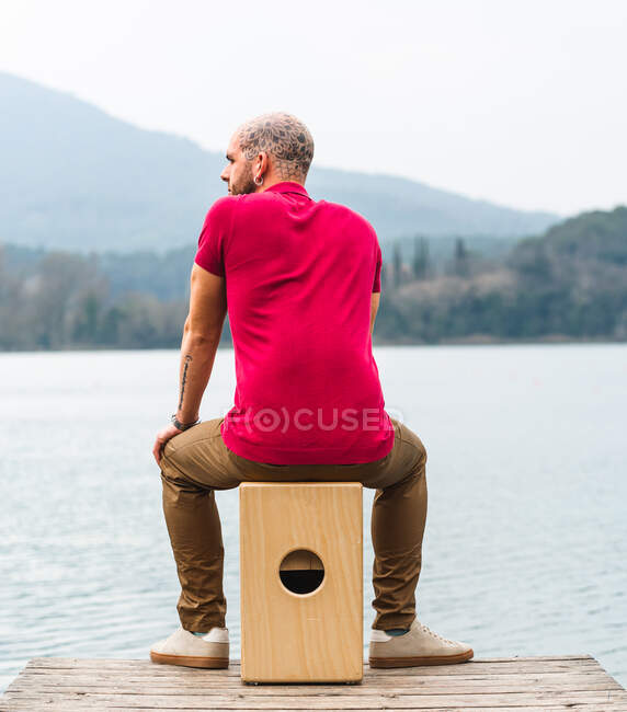 Homem percussionista concentrado sentado e jogando cajon no cais de madeira contra o rio calmo e montanhas em dia nublado — Fotografia de Stock