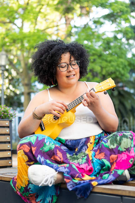 Cheerful female artist in ornamental clothes playing musical instrument on city bench — Stock Photo