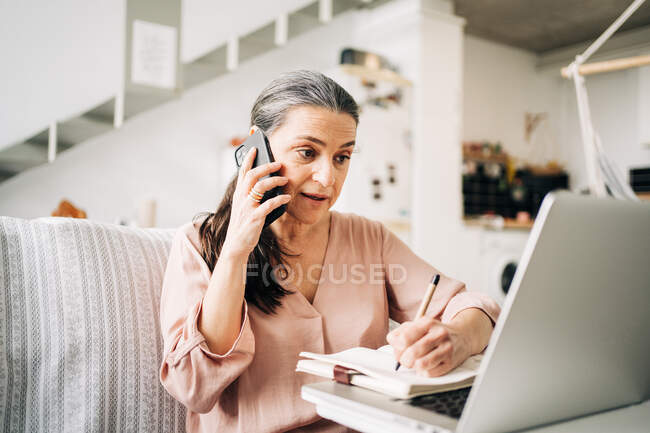 Concentrated middle aged female having phone call and taking notes in notepad while looking at screen of laptop in light room at home — Stock Photo
