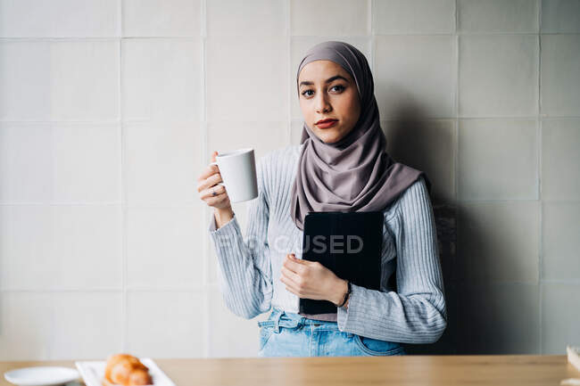 Retrato de mujer musulmana freelancer en pañuelo para la cabeza de pie en la mesa con una taza de bebida y una tableta mientras piensa en el proyecto y mira a la cámara - foto de stock