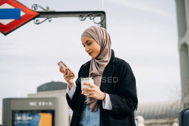 Deleitada mujer étnica en hijab y ropa de moda de pie con bebida para llevar mientras navega por Internet en el teléfono inteligente y disfruta del fin de semana en la ciudad - foto de stock