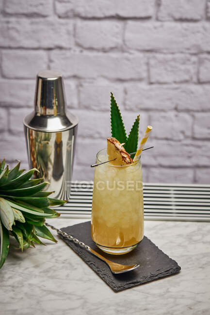 Cocktail giallo in vetro guarnito con pezzetto di ananas e foglie verdi con paglia di carta posta su sottobicchiere di ardesia con cucchiaio da bar — Foto stock