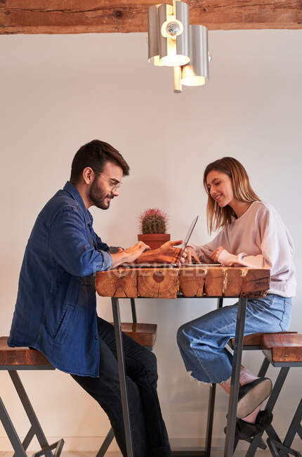 Side view of young Hispanic male freelancer working remotely on laptop sitting at table in kitchen with girlfriend browsing smartphone — Stock Photo