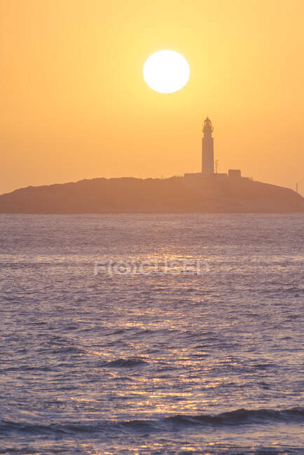 Paysage pittoresque d'eau ondulante de la côte de lavage de l'océan avec phare de grande hauteur placé à Faro de Trafalgar à Cadix en Espagne sous un ciel orange vif au lever du soleil — Photo de stock
