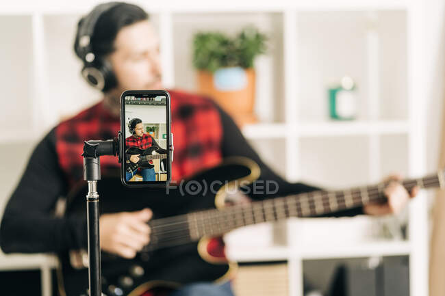Tripé com tela de celular representando fotografia de músico masculino em fone de ouvido tocando baixo em casa — Fotografia de Stock
