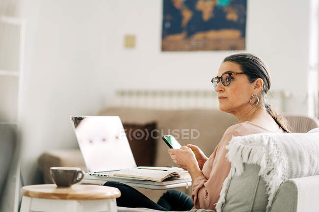 Vista lateral de la mujer de mediana edad concentrada que navega en el teléfono inteligente mientras trabaja en el ordenador portátil en la sala de luz en casa - foto de stock