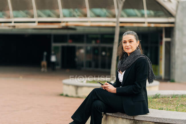 Seitenansicht einer emotionslosen weiblichen Textnachricht auf dem Handy, während sie auf einem Steinrand auf der Straße sitzt, mit Konstruktionen auf verschwommenem Hintergrund — Stockfoto