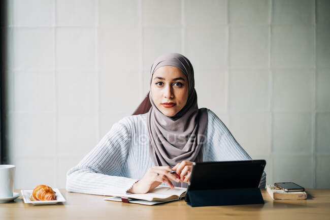 Задоволена мусульманка в Хіджабі працює зі своєю табличкою і сидить за столом у кафе, дивлячись на камеру. — стокове фото