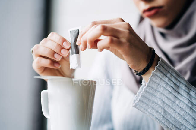 Crop anonymous Muslim female in hijab pouring sugar from paper packet in cup with hot drink in cafe — Stock Photo