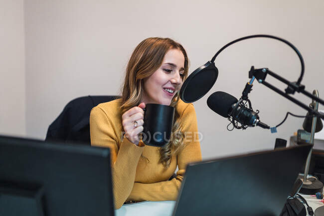 Mujer optimista sentada en la mesa con computadoras y bebiendo bebidas calientes mientras habla con el micrófono durante el trabajo en la moderna estación de radio - foto de stock