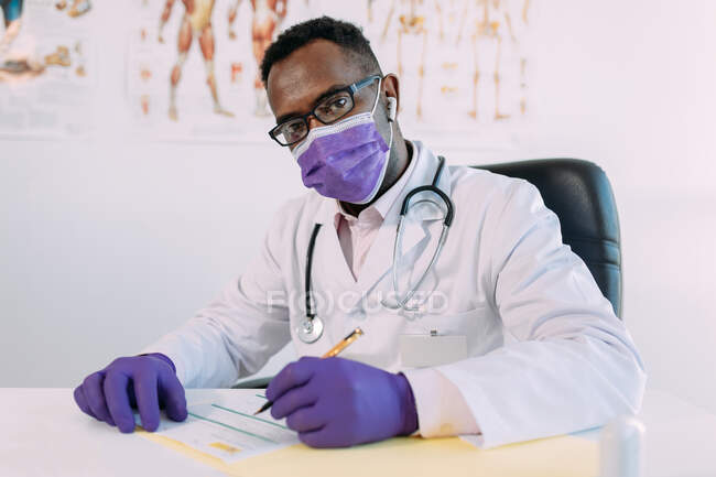 Médecin afro-américain en lunettes travaillant tout en écrivant dans le dossier du patient à table à l'hôpital — Photo de stock