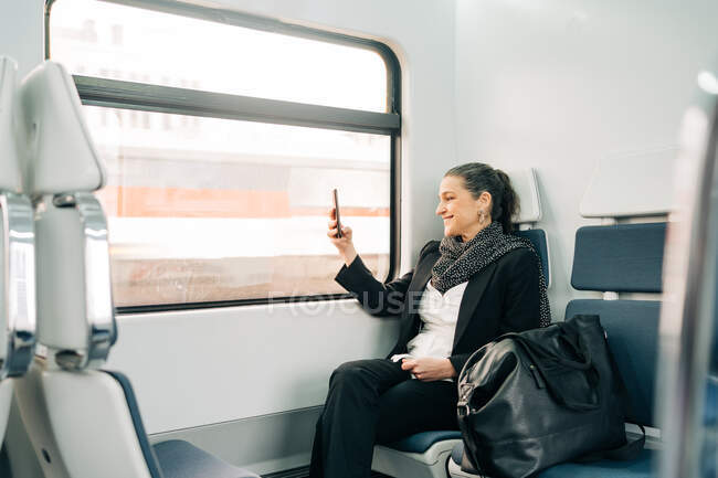 Vista lateral de la hembra de mediana edad positiva con bufanda tomando fotos en el teléfono celular mientras está sentada en el asiento del pasajero cerca de la ventana en el vagón durante el viaje - foto de stock