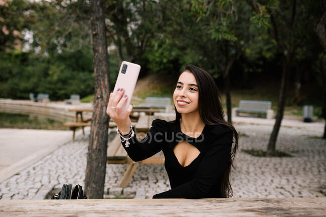 Empresaria con estilo positivo que toma autorretrato en el teléfono inteligente mientras está sentada en el banco de la ciudad - foto de stock