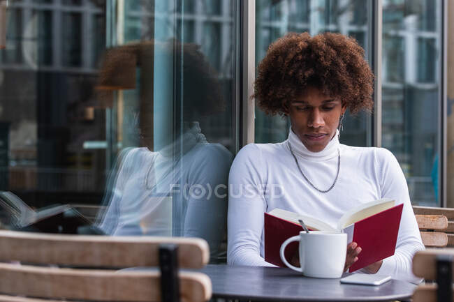 Спокійний молодий афроамериканець з кучерявим волоссям у модному одязі, що відпочиває на терасі кафе з чашкою кави і читає цікаву книжку — стокове фото
