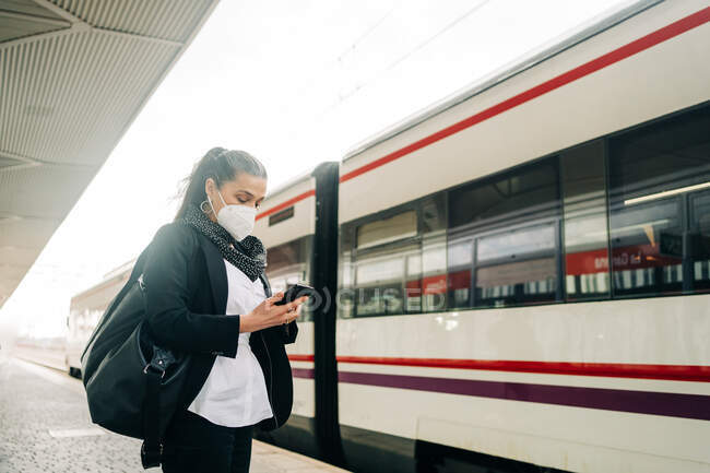 Grave femmina con borsa con respiratore protettivo messaggistica di testo sul cellulare mentre in piedi sulla stazione ferroviaria vicino al treno moderno durante il viaggio — Foto stock