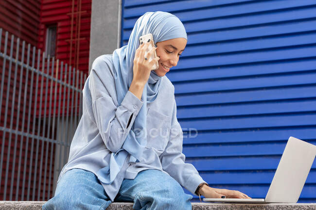 Smiling Muslim female in headscarf with netbook talking on cellphone in town — Stock Photo