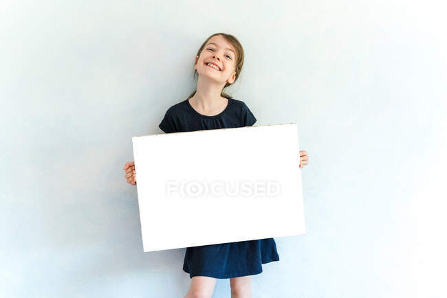 Menina feliz no desgaste casual mostrando tela vazia e olhando para a câmera com sorriso de dente enquanto está contra a parede branca — Fotografia de Stock
